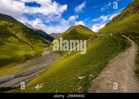 Ein schmaler Wanderpfad führt über einen steilen Grashang auf dem Weg von Girevi nach Chontio in der Munizipalität Akhmeta, Region Kacheti, Georgien. Unten links fließt ein Fluss durch ein breites Kiesbett. Grüne Bergrücken rahmen das Tal ein. Weiße Wolken ziehen über den tiefblauen Himmel. Der Weg von Girevi zum Atsunta-Pass folgt dem Kvakhidistskali-Tal und führt durch das verlassene Dorf Chontio. Chontio (georgisch ჭონთიო) liegt auf einer Höhe von etwa 2.200 Metern im Tuscheti-Nationalpark. Die Trekkingtour mit Packpferden umfasste fast 120 Kilometer und mehr als 5.000 Meter Auf- und Abstieg Stockfoto