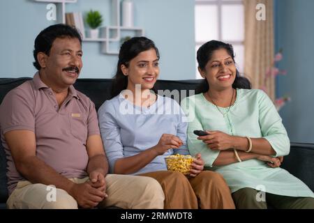 Entspannte glückliche indische Eltern mit Tochter, die fernsehen oder Fernsehen, indem sie zu Hause Popcorn isst - Konzept der familiären Bindung, Unterhaltung und Wochenende Stockfoto