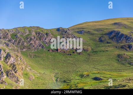 Ruinen von Steingebäuden liegen am Hang über Girevi in Tuscheti, in der Munizipalität Akhmeta, Region Kacheti, Georgien. Zwei Mauerreste ragen höher als der Rest. Links schneiden steil geneigte Schieferfelsen, die von Schluchten durchzogen sind, aus dem Hang heraus. Rechts ragen felsige Grate aus dem grasbewachsenen Hang. Unten fällt eine grüne Wiese zum Tal ab. Die Morgensonne erhellt den Hang unter einem blauen Himmel. Girevi (georgisch გირევი) liegt auf einer Höhe von etwa 2.060 Metern am Fluss Pirikita Alazani. Das Dorf liegt innerhalb eines geschützten Gebiets. Einige Familien kehren im Sommer zurück, aber im Winter ist es verlassen Stockfoto