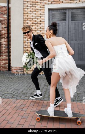 afroamerikanische Frau im Hochzeitskleid reitet Longboard in der Nähe eines fröhlichen Bräutigams in einer Sonnenbrille Stockfoto