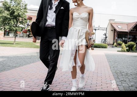 Kurzer Blick auf elegante multiethnische Paare, die mit Longboard und Skateboard spazieren gehen, Hochzeit in der Stadt Stockfoto