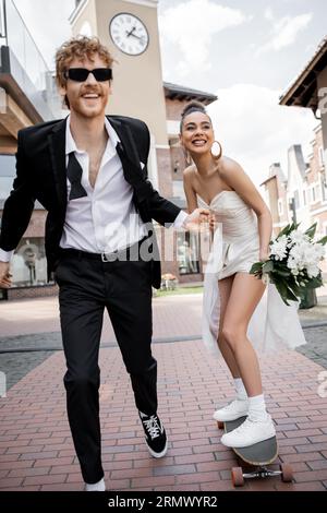 Elegante afroamerikanische Frau in Hochzeitskleid reitet Longboard in der Nähe des Bräutigams in der Sonnenbrille Stockfoto