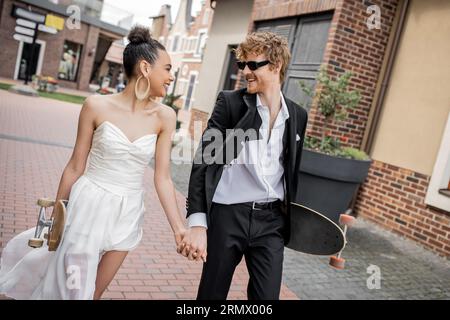 Glückliches interrassisches Paar mit Longboard und Skateboard auf der Straße zu Fuß, Hochzeitskleidung, Sonnenbrille Stockfoto