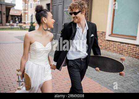 Multiethnische Paare gehen mit Longboard und Skateboard auf der Straße, Hochzeitsoutfit, Sonnenbrille Stockfoto