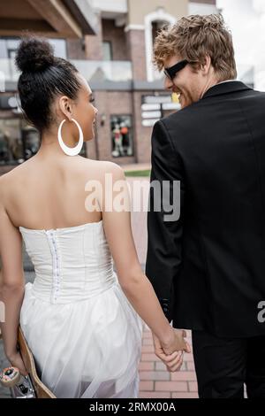 Rothaariger Bräutigam in Sonnenbrille, afroamerikanische Braut mit Longboard, die sich auf der Straße ansieht Stockfoto