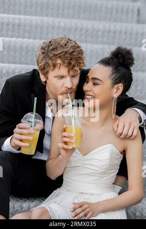 Rothaariger Bräutigam, der Orangensaft aus Stroh in der Nähe einer fröhlichen amerikanischen Braut trinkt, Hochzeit in der Stadt, Spaß Stockfoto