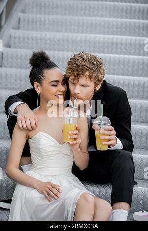 Hochzeit in der Stadt, afroamerikanische Braut lächelt in der Nähe des rothaarigen Bräutigams und trinkt Orangensaft aus dem Stroh Stockfoto