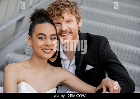 Porträt von sorglosen und stilvollen, interrassischen Brautpaaren, die vor der Kamera lächeln, Hochzeit im Freien Stockfoto