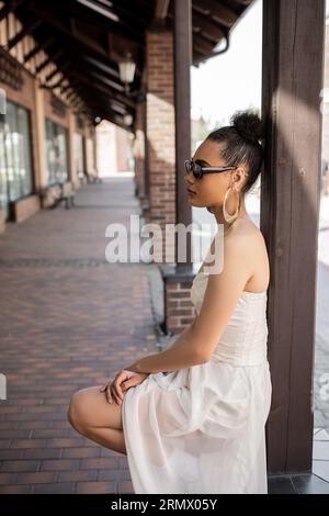 Seitenansicht der bezaubernden afroamerikanischen Frau in Hochzeitskleid und Sonnenbrille auf der urbanen Straße Stockfoto