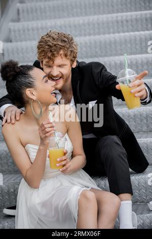 Fröhlicher rothaariger Bräutigam, der mit dem Finger auf die afroamerikanische Braut zeigt, mit Orangensaft auf der Straße Stockfoto
