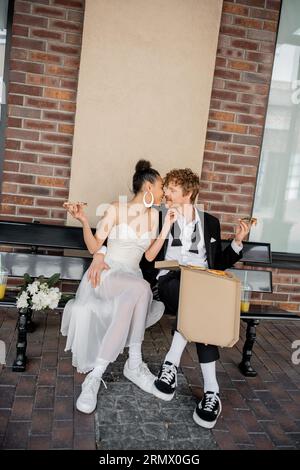 Elegantes, multirassisches Paar mit Pizzakuss auf der Bank, Hochzeit in der Stadt, Feier im Freien Stockfoto