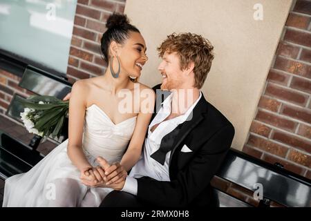 Ein junges multiethnisches Paar lächelt einander an und hält die Hände an Bank, Hochzeit, städtisches Setting Stockfoto