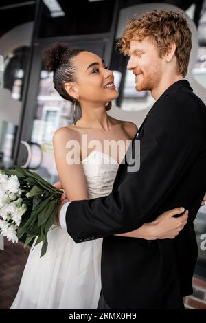 Überfreuliches und elegantes Paar aus verschiedenen Rassen in Hochzeitskleidung, das sich auf der Straße der Stadt umgibt Stockfoto