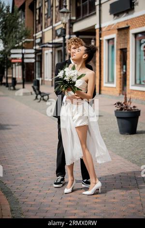 Junger rothaariger Mann, der afroamerikanische Frau in Hochzeitskleid umarmt, Blumen, Freude, Hochzeit in der Stadt Stockfoto