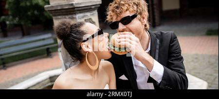 Multiethnisches frisch vermähltes Paar in Sonnenbrille, das Burger beißt, draußen Spaß hat, Banner Stockfoto