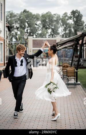 Hochzeit in der modernen Stadt, elegantes multiethnisches Paar, das Hände hält und auf der Straße tanzt, Glück Stockfoto