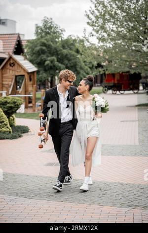 Glückliches und stilvolles multiethnisches Paar, das mit Longboard und Blumen spaziert und sich gegenseitig anlächelt Stockfoto