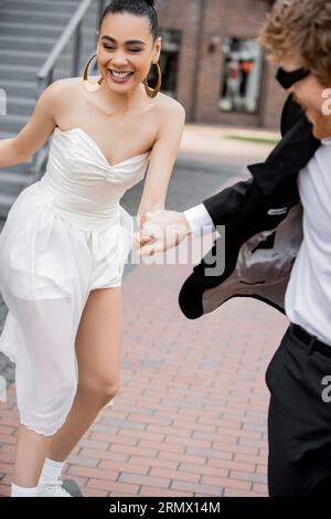 unbekümmerte afroamerikanische Frau im Hochzeitskleid, die Hände mit dem Bräutigam hält und in der Stadt läuft Stockfoto