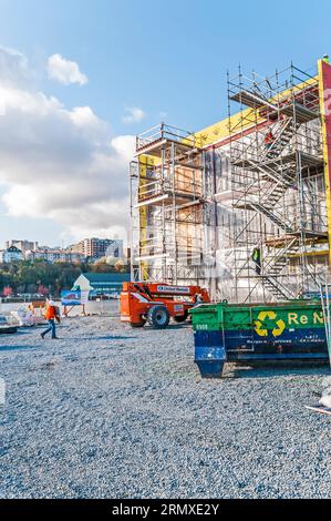 Ein orangefarbener Miethochstuhl mit einem Arm, der bis zum Gebäudedach auf einer Baustelle reicht, während ein Bauarbeiter in einem Schutzhelm auf der Baustelle spaziert. Stockfoto