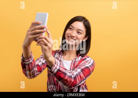 Hübsche lächelnde asiatische Dame mit stilvollem, legeren Outfit, die ihr Handy hält und Selfie macht, gelber Studiohintergrund Stockfoto