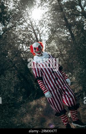 Ein verrückter böser Rothaariger Clown, der ein weiß-rot gestreiftes Kostüm mit einem weißen Fell trägt, steht im Wald und starrt den Betrachter mit einem gruseligen Lächeln an Stockfoto