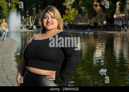 Junge blonde argentinierin plus Größe Latina Frau im öffentlichen Park mit Lagune im Hintergrund, sie steht posierend mit den Händen auf ihrer Taille glücklich smi Stockfoto