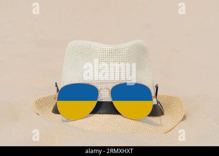 Sonnenbrillen mit Gläsern in Form einer Flagge der Ukraine und einem Hut liegen auf Sand. Konzept von Sommerferien und Reisen in der Ukraine. Sommerruhe Stockfoto