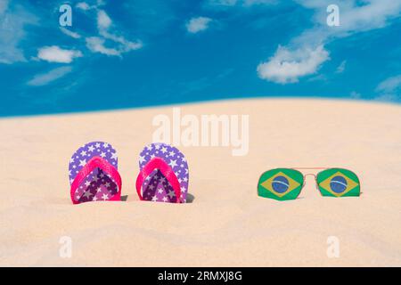 Sonnenbrillen mit Brillen in Form einer brasilianischen Flagge und Flip-Flops liegen auf Sand am blauen Himmel. Konzept von Sommerferien und Reisen in Brasilien Stockfoto