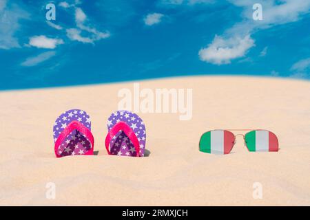 Sonnenbrillen mit Gläsern in Form einer italienischen Flagge und Flip-Flops liegen auf Sand gegen blauen Himmel. Konzept der Sommerferien und Reisen in Italien Stockfoto