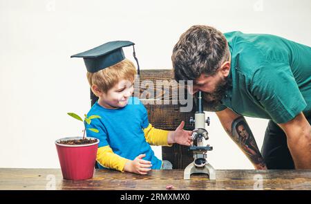 Biologiestunden in der Grundschule. Kleiner Junge im Graduiertenhut und bärtiger Professor, der mit dem Mikroskop arbeitet. Lehrer mit einem kleinen Jungen in der Schule Stockfoto