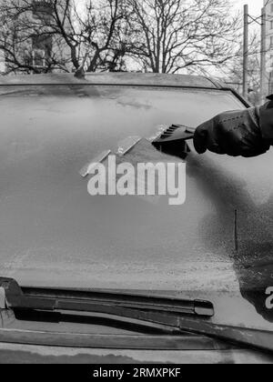 Reinigung der Autoscheiben im Winter. Man kratzt Raureif mit einem Plastikkratzer von der Windschutzscheibe eines Autos. Tragen Sie einen schwarzen Lederhandschuh in der Kälte. Stockfoto