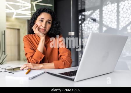 Eine müde und nachdenkliche junge Frau sitzt im Büro am Tisch und schaut auf den Laptop-Bildschirm, wobei ihr Kopf träumerisch auf der Hand liegt. Stockfoto