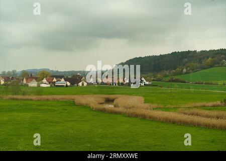 Über dem deutschen Dorf gibt es Regenwolken, neben dem Dorf gibt es einen Wald, ein Feld und einen Teich. Stockfoto