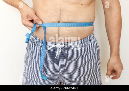 Der Mann misst seinen dicken Bauch mit Maßband und zeigt Daumen nach oben. Konzept der Gewichtsabnahme, Gesundheitsprobleme von übergewichtigen Menschen. Nahrungskontrolle und Akt Stockfoto