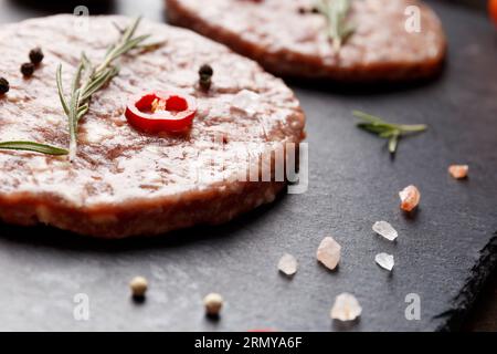 Rohes, frisches Hackfleisch, Burger-Patties, Rosmarin, Gemüse und Gewürze auf schwarzem Steinteller auf dunklem Hintergrund. Zutaten für hausgemachten Hamburger. Stockfoto