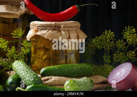 Eingelegte Gurken in einem Glas, Gurkensalat in Dosen, frische Gurken, Kräuter und Gewürze auf dunklem Hintergrund, Nahaufnahme. Stockfoto