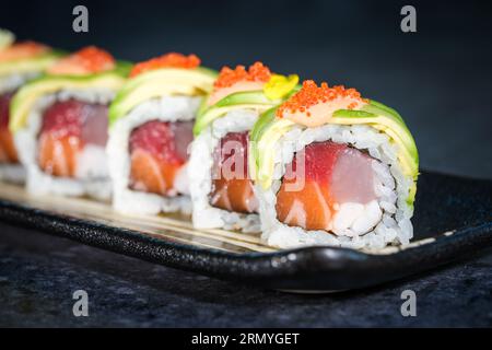 Nahaufnahme von appetitlichem Tobiko-Reh mit frischen grünen Avocadoscheiben auf uramakifarbenen Sushi-Brötchen, serviert auf Tablett auf Marmortheke Stockfoto