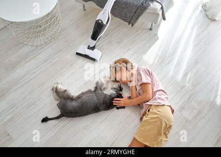 Von oben Junge in legerer Kleidung streichelte Schnauzer Hund, während auf dem Boden in der Nähe des Staubsaugers während der Hausroutine liegen Stockfoto