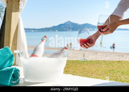 Crop anonyme Person Putting Paar Becher mit Rosenchampagner auf Himmelbett in der Nähe des Meeres während romantische Datum am Strand am Sommertag platziert Stockfoto