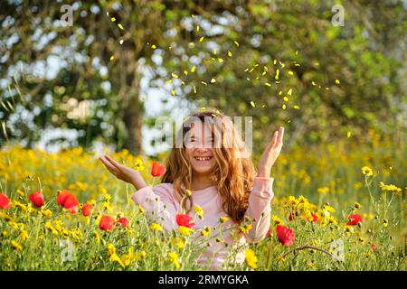 Optimistisches Mädchen lächelt und wirft Blütenblätter in die Luft, während sie sich in blühender Wiese am sonnigen Tag entspannt Stockfoto