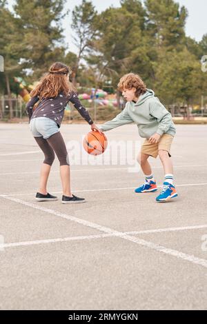 Freundliches Teenager-Mädchen und -Junge spielen Basketball auf Asphalt-Spielplatz, während sie im Sommer zusammen unterhalten Stockfoto