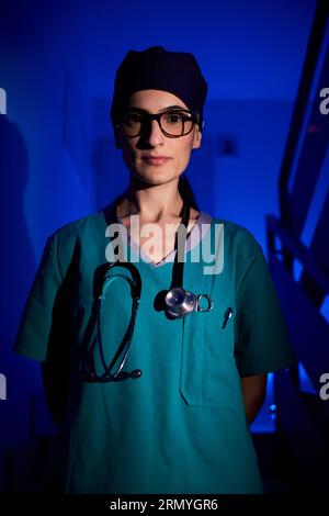 Selbstbewusste Erwachsene Frau in medizinischer Uniform mit Stethoskop, das auf Treppen mit den Händen hinter dem Rücken steht und die Kamera im dunklen Raum unter blauem Licht betrachtet Stockfoto