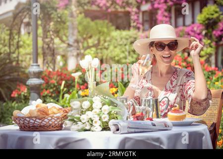 Stockfoto einer glücklichen Frau, die eine Sonnenbrille trägt und im Garten ein Glas Champagner trinkt. Stockfoto