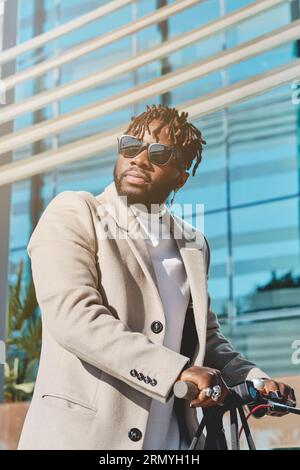 Nahaufnahme eines selbstbewussten bärtigen afroamerikanischen Mannes, der Sonnenbrille und Mantel trägt, der gegen ein modernes Gebäude steht, während er mit Roller und Lookin steht Stockfoto