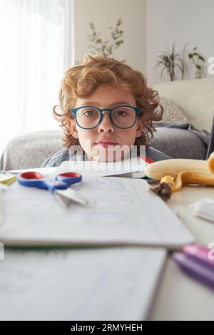 Junge mit Brille und lockigem Haar, der in die Kamera schaut, während er sich auf ein Notebook auf dem Tisch lehnt und zu Hause Hausaufgaben macht Stockfoto