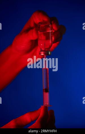 Nicht erkennbare Person, die die mit COVID 19-Impfstoff gefüllte Spritze und Durchstechflasche vorführt und sich auf die Injektion in einem dunklen Raum auf blauem Hintergrund vorbereitet Stockfoto
