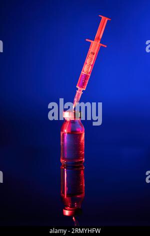 Medizinische Spritze mit Nadel, die in die Durchstechflasche aus Impfglas in rotem Licht eingeschoben wird, das auf der Oberfläche vor blauem Hintergrund reflektiert Stockfoto