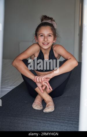 Voller Körper eines positiven jungen Mädchens mit Haarknospen in Aktivkleidung und Turnschuhen, die auf dem Bett mit überkreuzten Beinen sitzen und nach vorne schauen Stockfoto