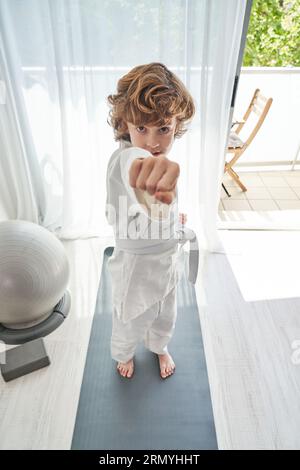 Von oben ein Kind im Kimono mit weißem Gürtel, der auf einer grauen Sportmatte steht und mit geballter Faust auf die Kamera zeigt, während er im Wohnzimmer zu Hause Judo macht Stockfoto