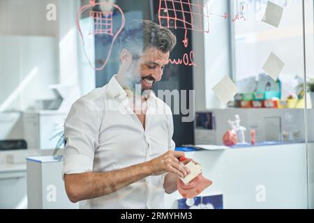 Durch das Fenster eines männlichen Ingenieurs, der sich in formeller Kleidung mit Atemschutzmaske trägt und während er in der Nähe einer Glaswand steht, Haftnotizen mit schriftlichen Informationen macht Stockfoto